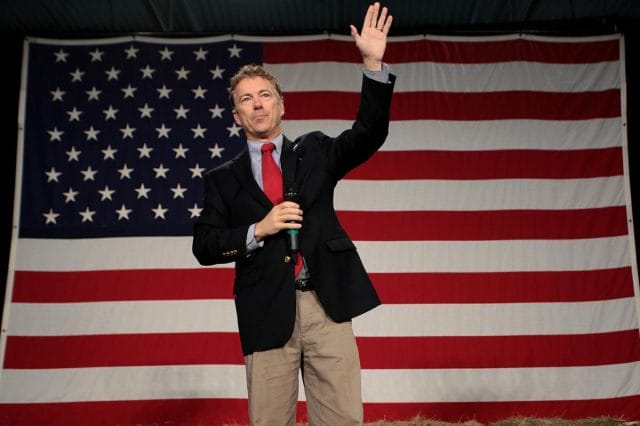 Rand Paul in a suit and tie holding the hand up in front of a flag