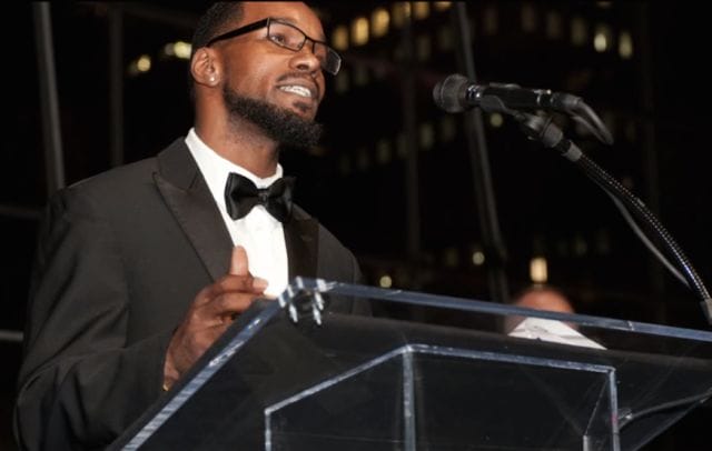 Kevin Berthia in a tuxedo smiling at a podium