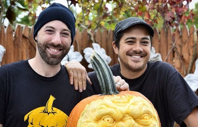 two men wearing black shirts and holding a carved pumpkin