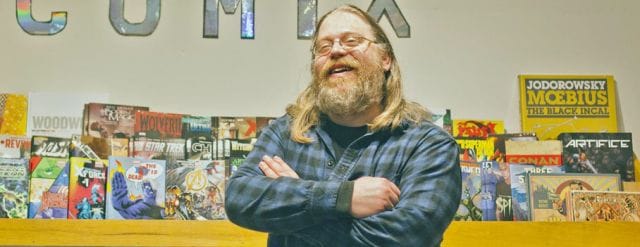 a man with a beard sitting at a desk with his arms crossed