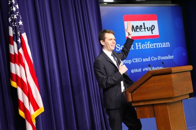 Scott Heiferman in a suit standing at a podium with a microphone and a flag behind him