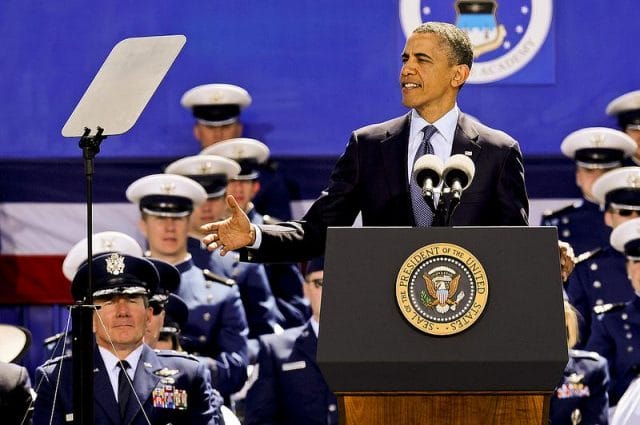 a person in a suit and tie speaking into a microphone in front of Barack Obama et al. in