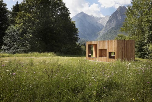 a wooden building in a grassy field with mountains in the background