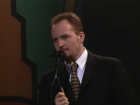 Louis C.K. in a suit and tie speaking into a microphone